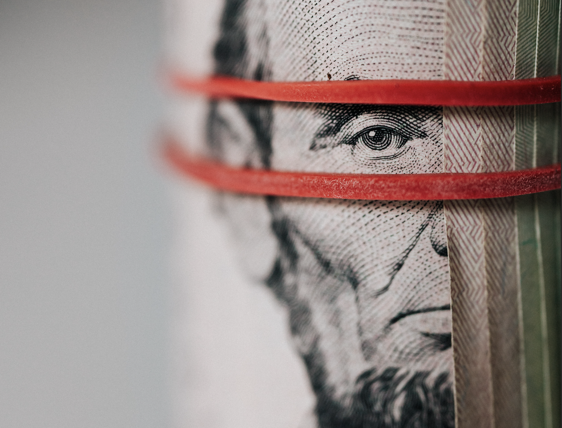 Closeup view of a roll of five-dollar bills held together by a red rubber band that frames Abraham Lincoln's eyes, as if he is watching the viewer.